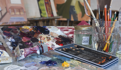 taiteilijan pöydällä lyijykyniä ja maalauksia. Pöydällä lasipurkissa värikyniä ja maalauspensseleitä. Taustalla maalauksia.