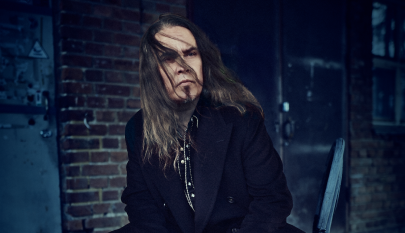 Pitkähiuksinen henkilö tummassa takissa istuu teollisessa ympäristössä tiiliseinän edessä. Kuvan tunnelma on tumma ja voimakas.