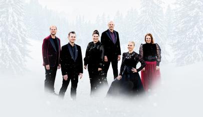 Kuusi esiintyjää seisoo lumisessa metsämaisemassa talvisissa juhla-asuissa. Taustalla näkyy lumisia kuusia ja kuvan alareunassa vaalea lumisumu.
