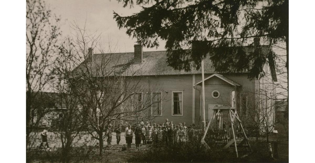 Kansakoulu taustalla, lapsiryhmä pihalla katse suunnattuna kameraan. Etualalla puita ja pensaita sekä vastakkain istuttava keinu.