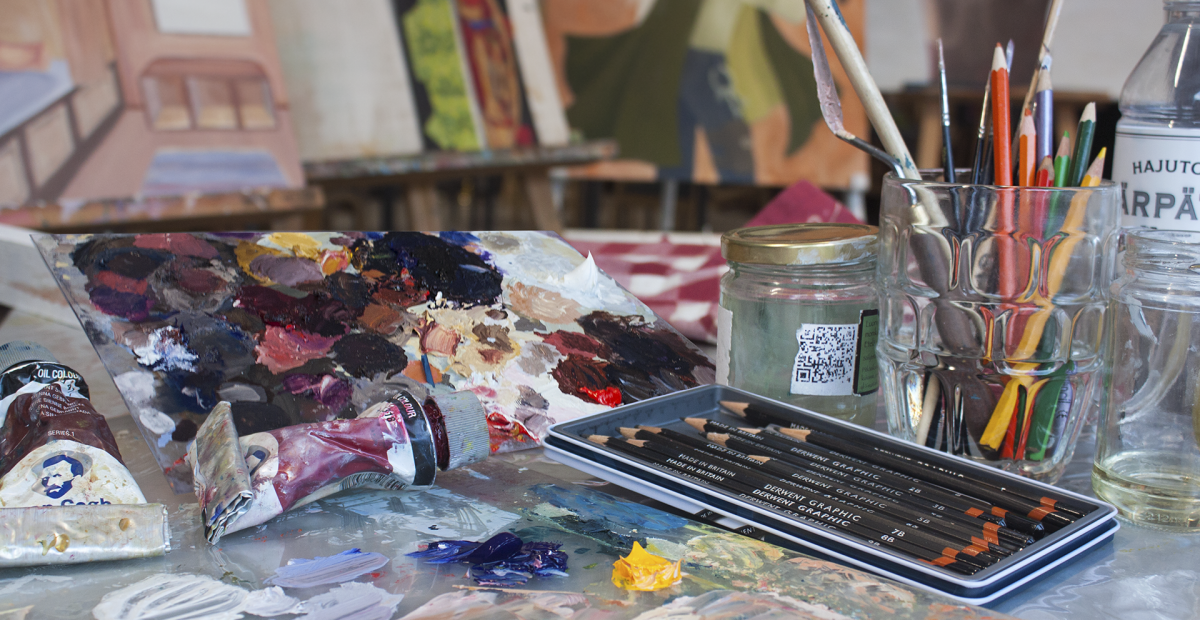 taiteilijan pöydällä lyijykyniä ja maalauksia. Pöydällä lasipurkissa värikyniä ja maalauspensseleitä. Taustalla maalauksia.